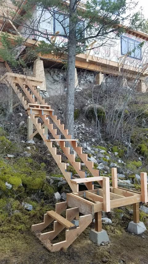 Wooden outdoor staircase and bridge-like structure on a rocky, mossy hillside by a house.