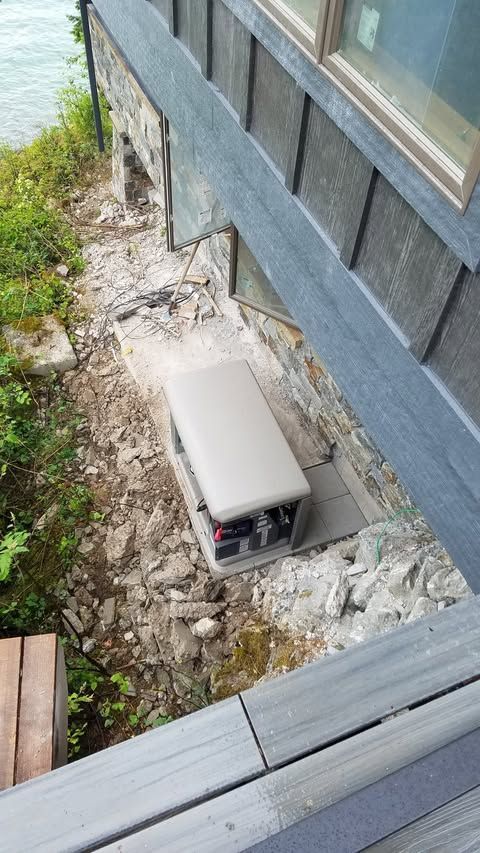 Small white utility box beside a gray house, set in a narrow rocky strip near the water.