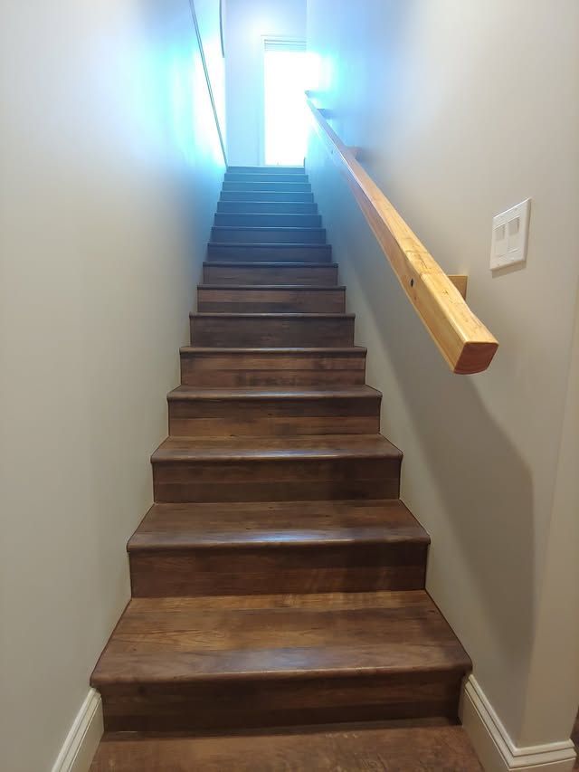 Wooden staircase in a narrow hallway leading up to a bright doorway, with a handrail on the right.