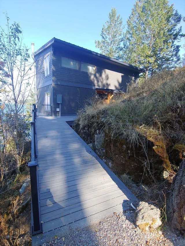 Wooden ramp leading to a modern dark house on a wooded hillside under bright sun
