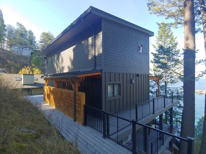 Modern two-story dark wood house on a hillside with a sloped walkway and trees around it