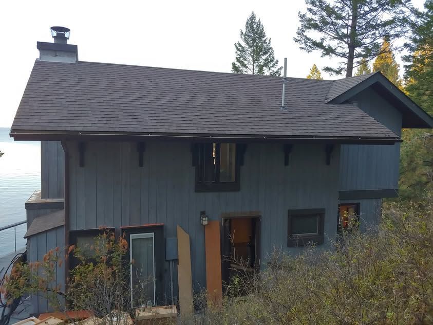 Dark gray lakeside house with steep roof, surrounded by trees and shrubs