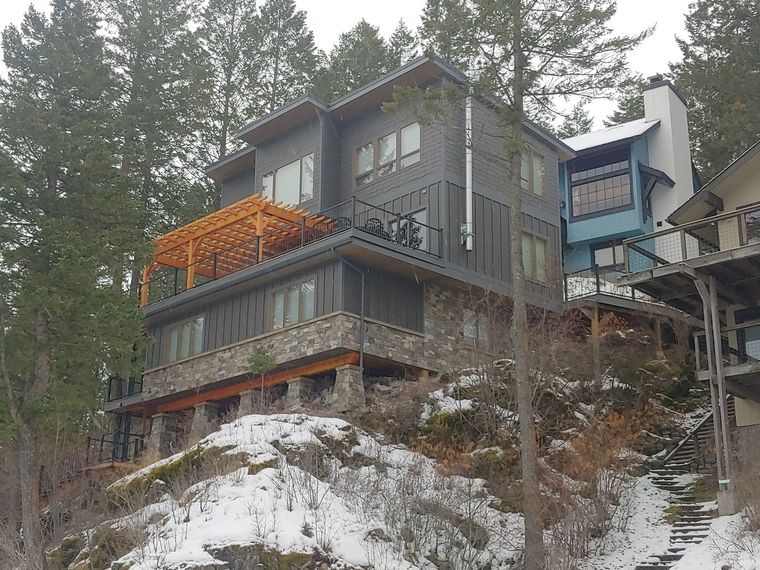 Snowy hillside house with dark gray siding, orange deck, and evergreen trees in the background