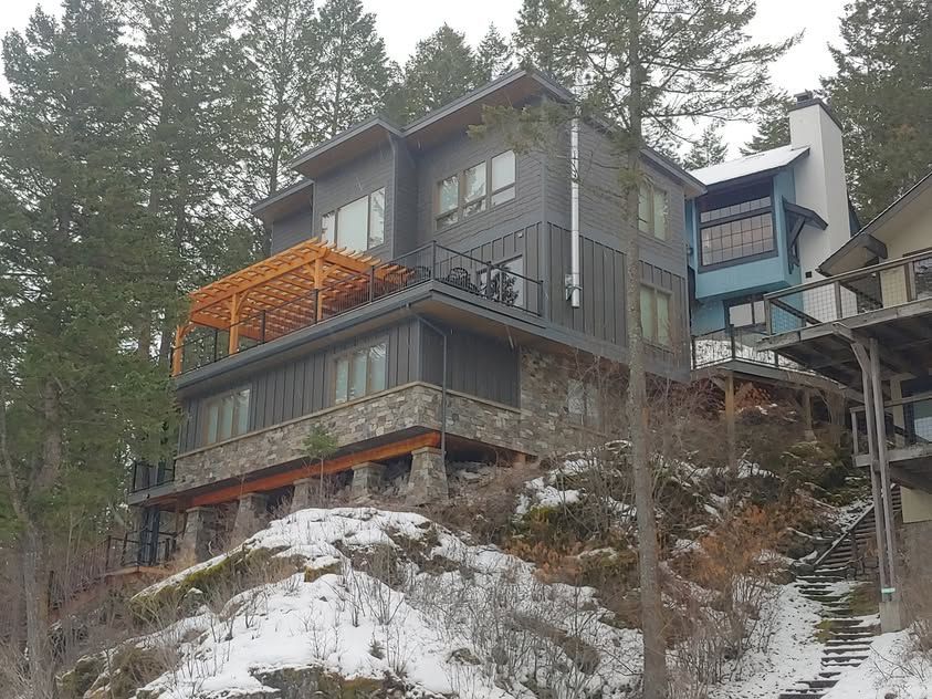 Modern hillside house with dark siding, orange balcony, and snowy ground among pine trees