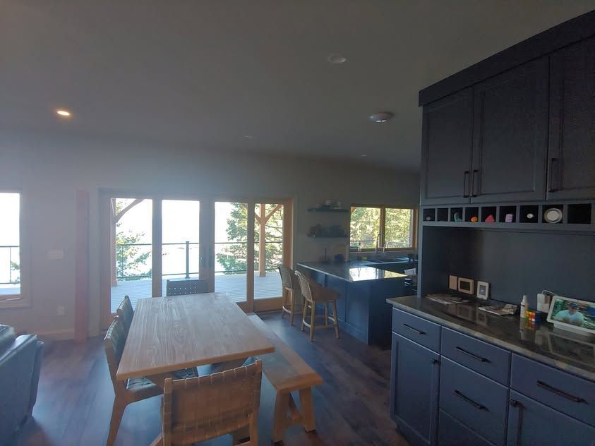 Open kitchen and dining area with dark cabinets, long wooden table, and large windows overlooking trees