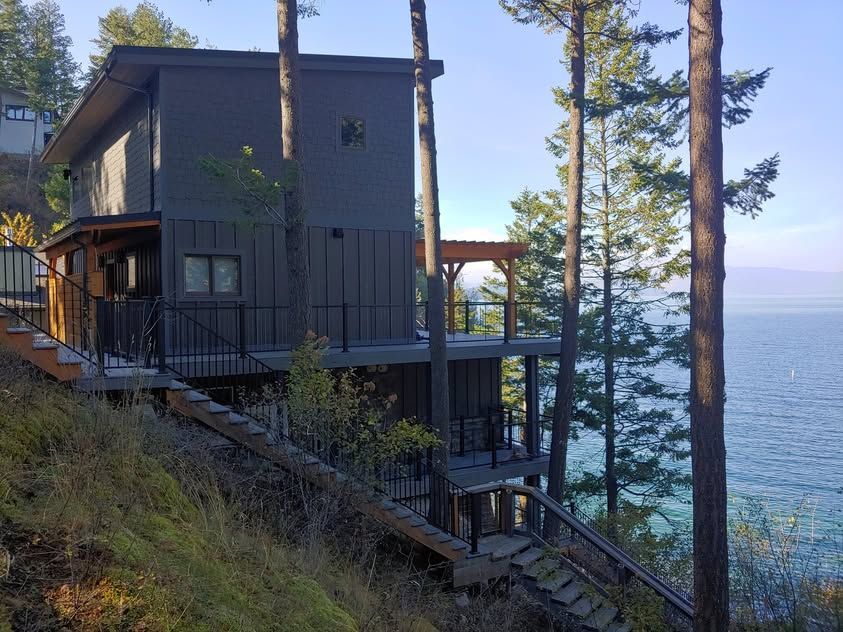 Modern gray lakeside house on a wooded hillside with stairs and tall pine trees overlooking the water