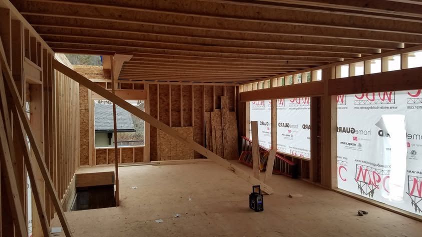 Interior of a house under construction with exposed wooden framing, unfinished floors, and large windows.