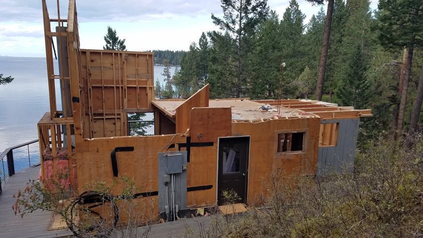 Rustic wooden cabin under construction by a lake, with tools and a forested shoreline behind it.