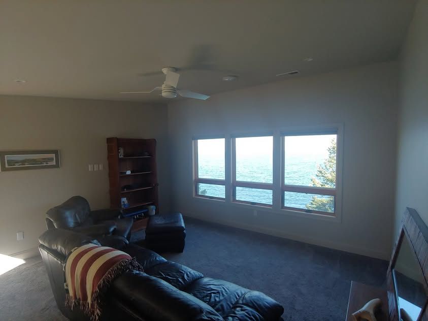Living room with black leather sofas, ceiling fan, bookshelf, and bright windows overlooking outdoors
