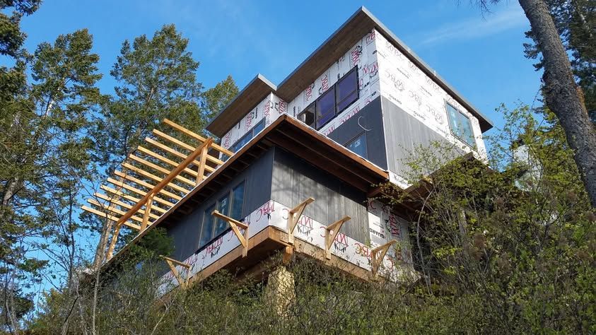Modern house with white and brown siding on a wooded hillside, viewed from below under a blue sky