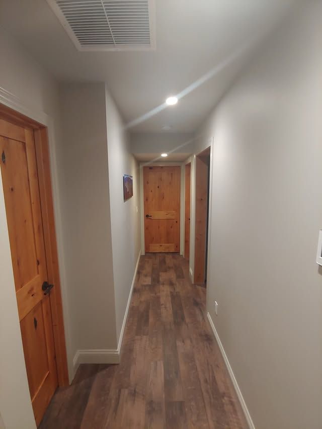 Narrow hallway with wood floors, white walls, ceiling lights, and wooden doors at both ends.