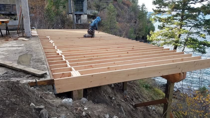 Wooden deck frame under construction beside a lakeside cabin, with a worker crouching on the joists.