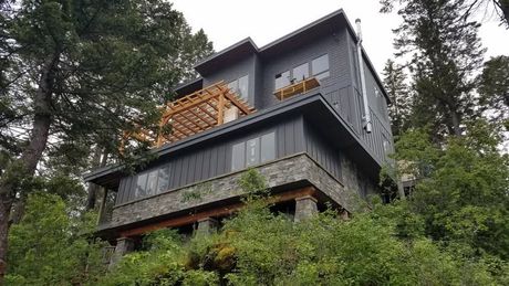 Modern dark-gray house with wooden balconies nestled among tall trees on a forested hillside