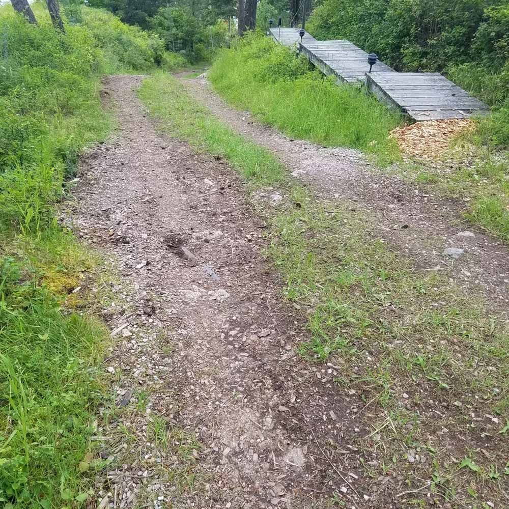 Muddy dirt road through lush green vegetation beside stacked metal sheets on the right