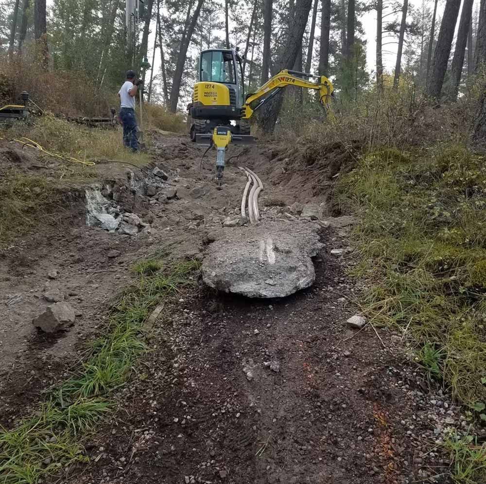 Excavator clearing a muddy forest trail with a worker nearby and large rocks in the path