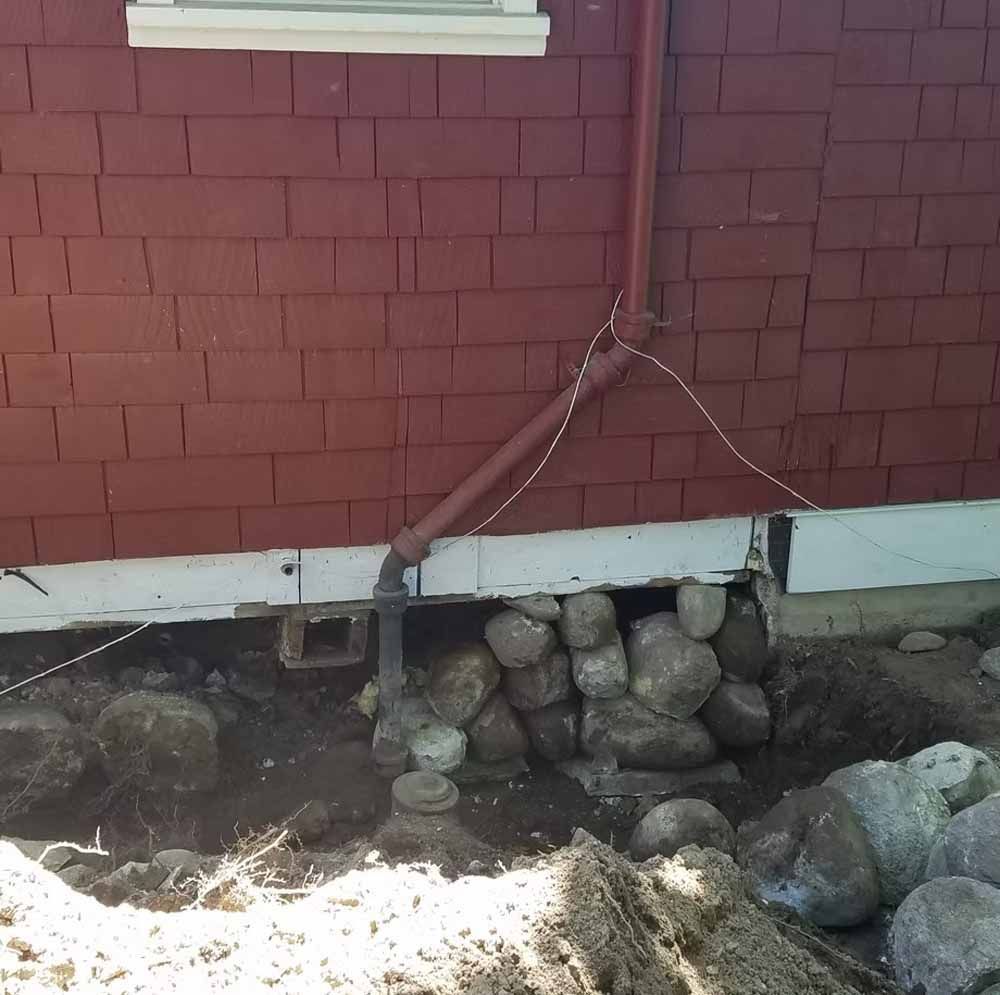 Red house foundation with downspout and rocks piled along the base next to a trench.