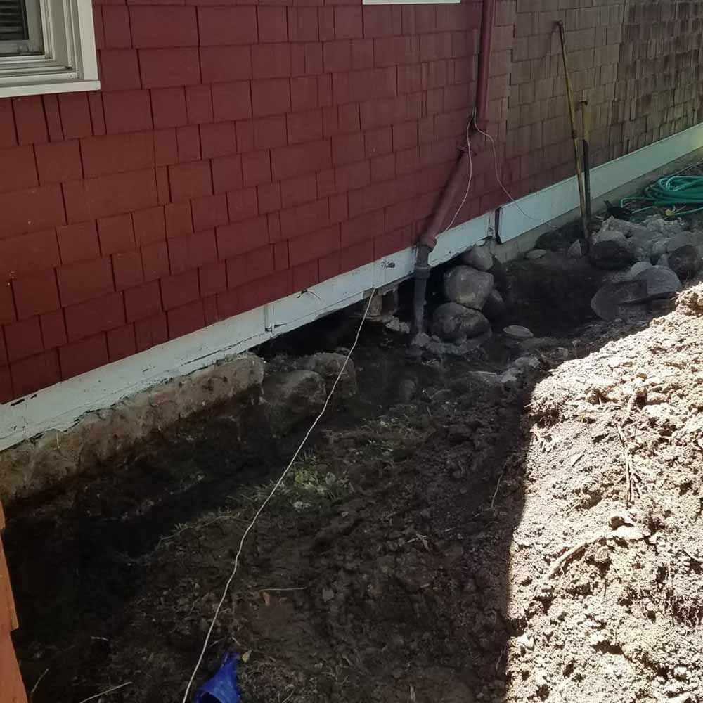 House foundation with exposed soil, rocks, and a sagging cable beside a red exterior wall
