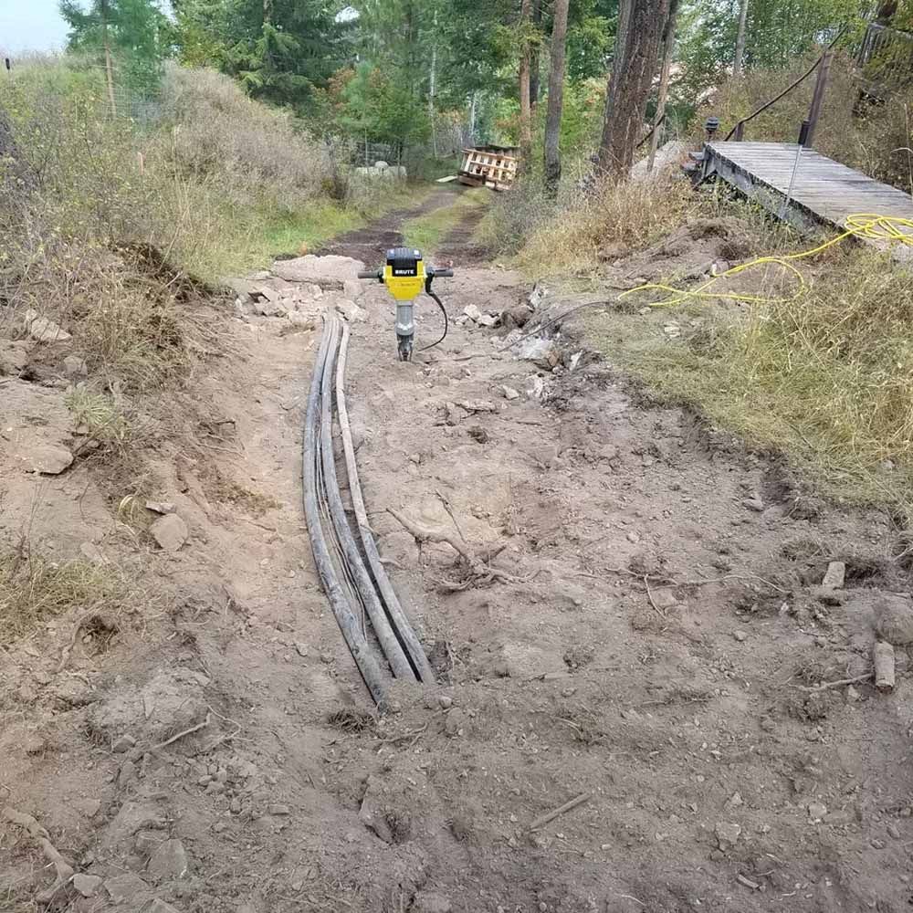 Dirt construction trench with cables, a yellow survey marker, and a small wooden shed beside a wooded path