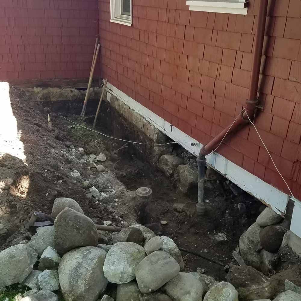 Rock-bordered dirt trench beside a red brick house with exposed pipes and a small window