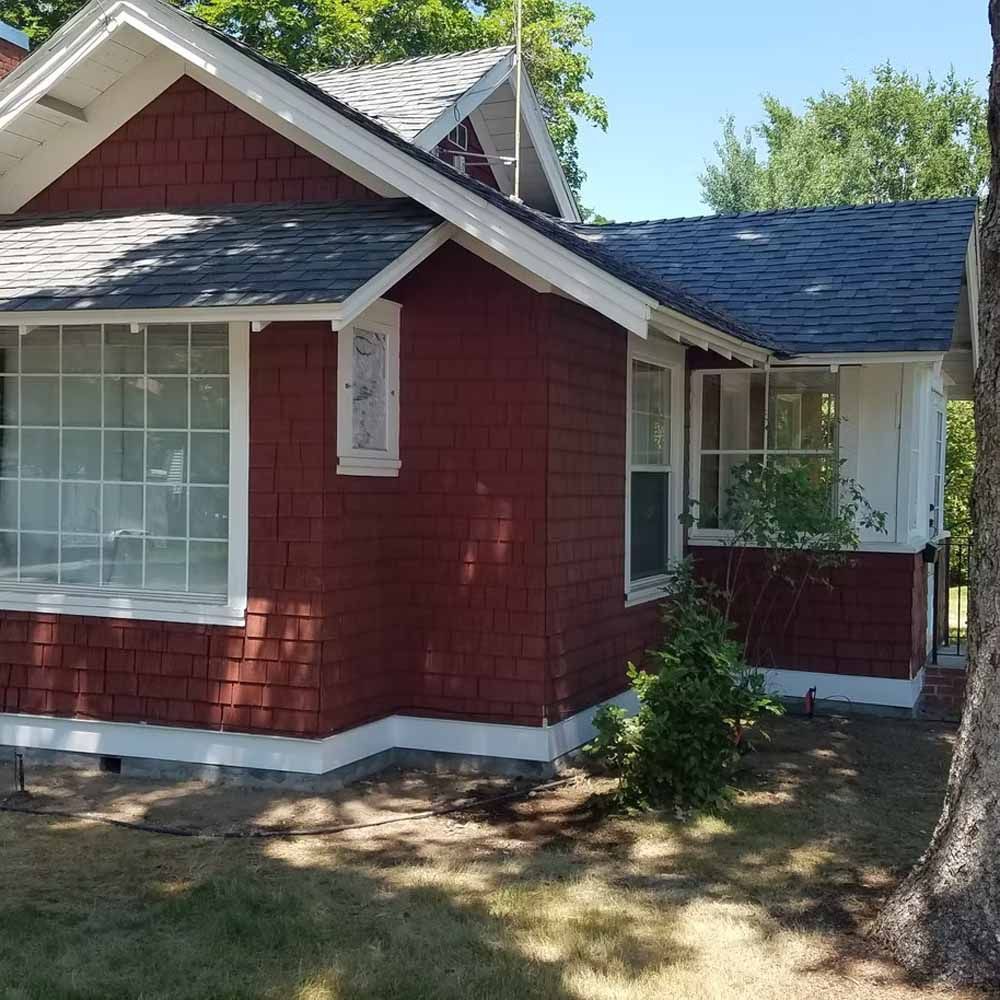 Red shingle house with white trim, large front window, and small porch under trees.