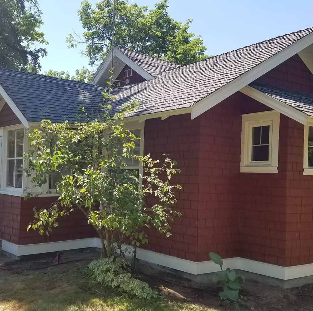 Red house with gray shingle roof and white trim, partly shaded by a tree in a sunny yard