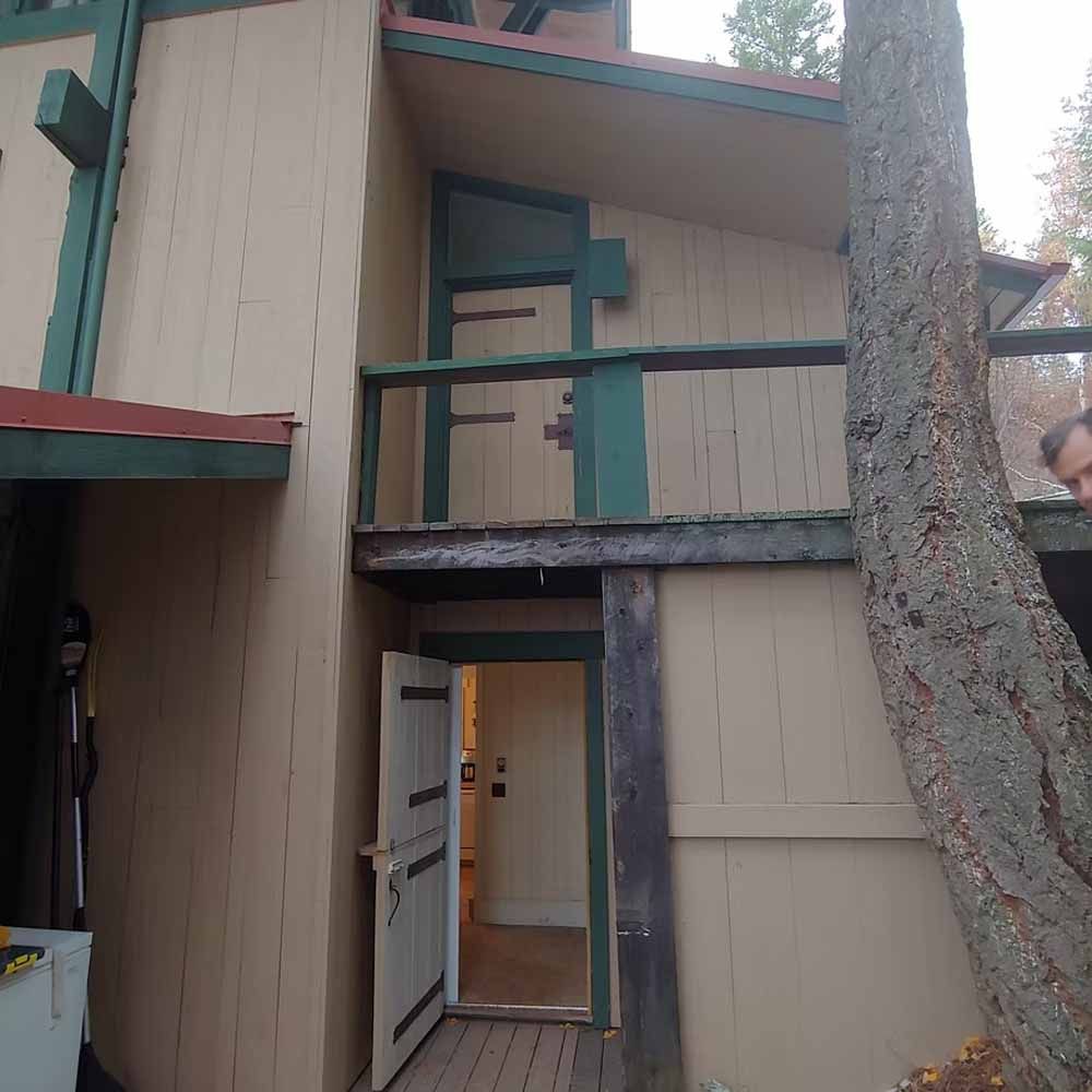 Two-story beige apartment exterior with open white door, green trim, and a large tree in front.