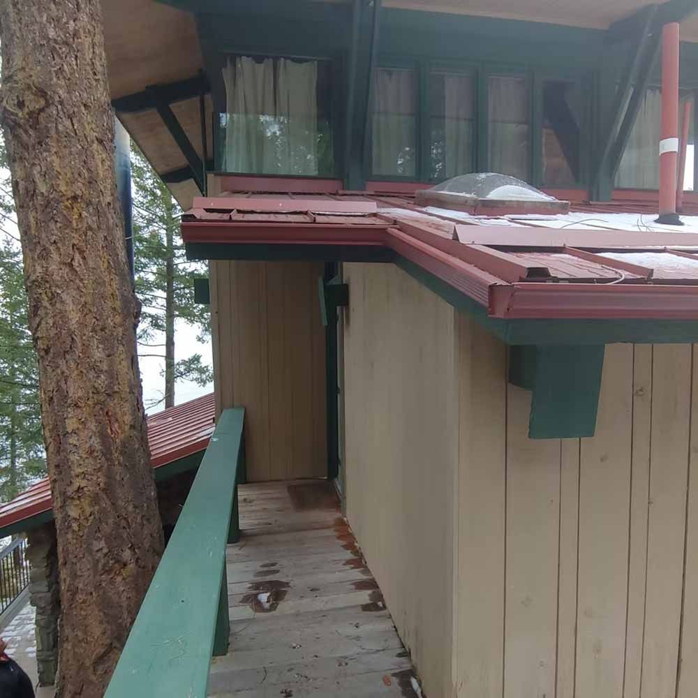 Narrow wooden walkway beside a beige cabin with red roof, surrounded by trees in a forest setting