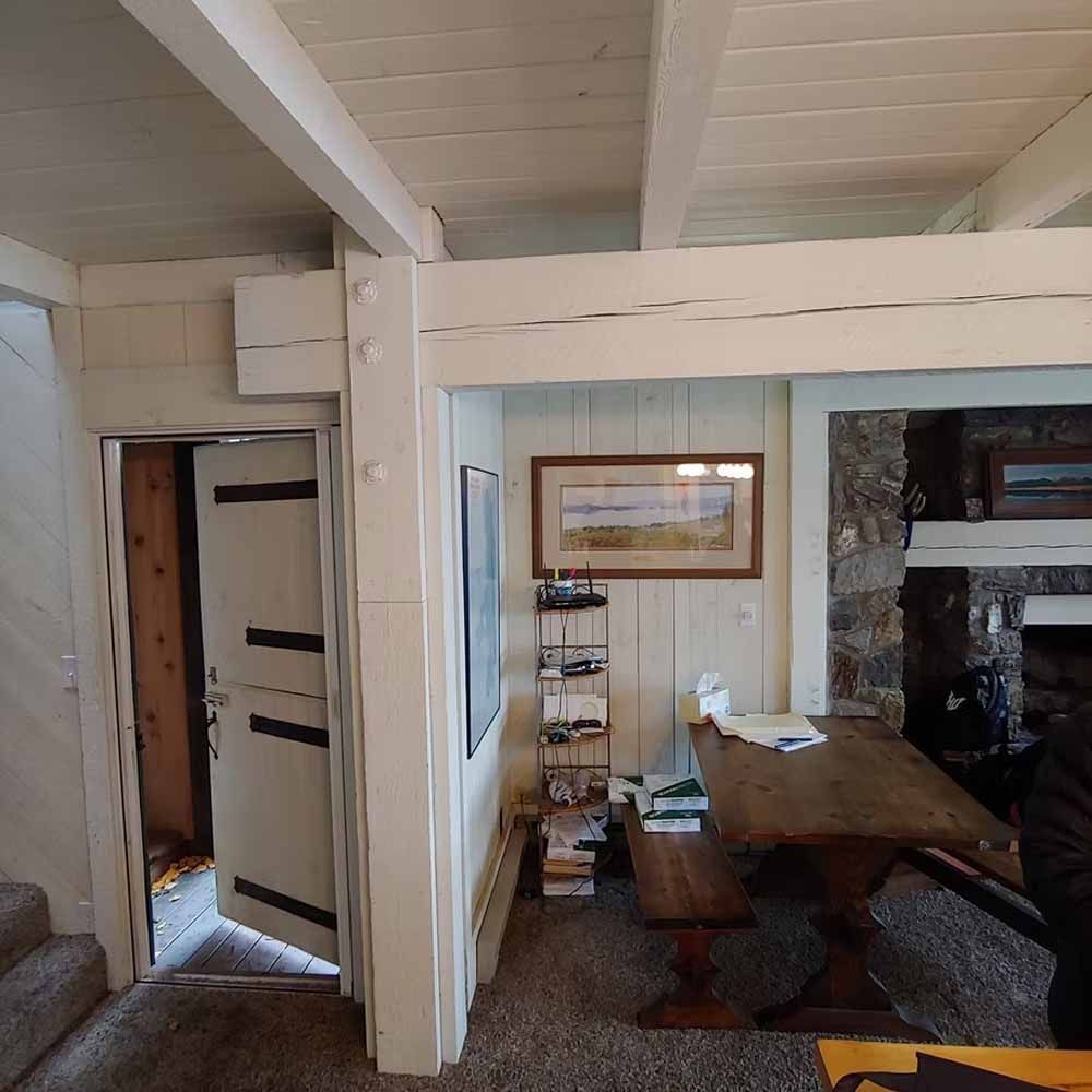Rustic cabin dining area with wooden table, bench, stone fireplace, and open doorway to another room.