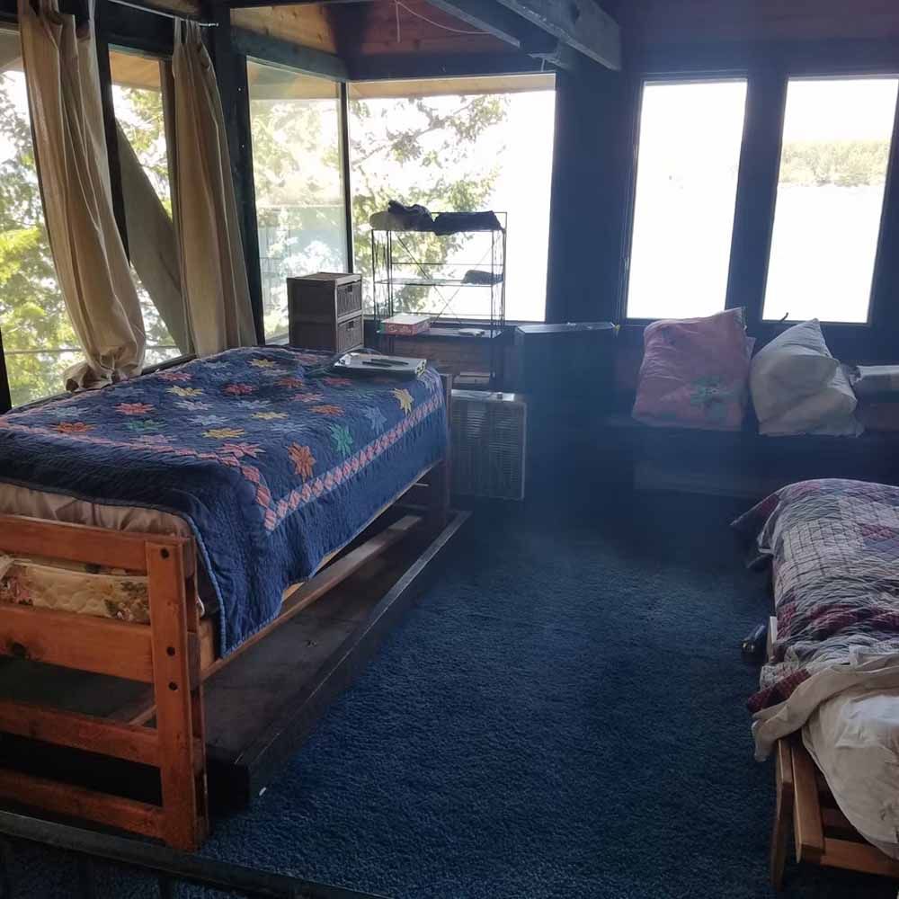 Rustic cabin bedroom with two wooden beds, blue rug, and large windows overlooking bright water.