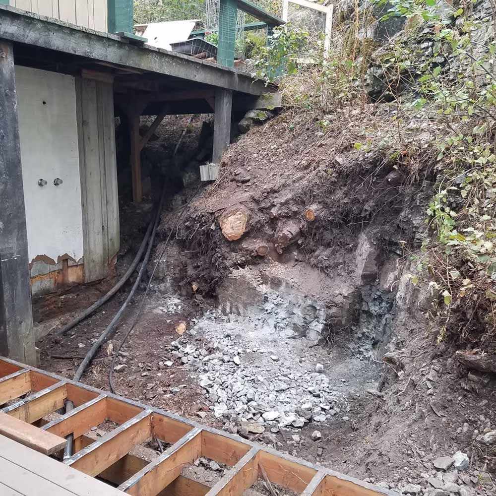 Collapsed dirt slope beside a building, exposing cables and debris under a concrete overhang.