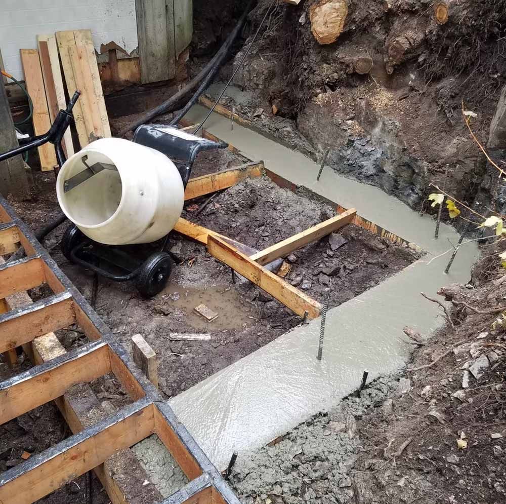 Concrete foundation formwork with rebar, a cement mixer, and wooden supports in a dug-out construction site.