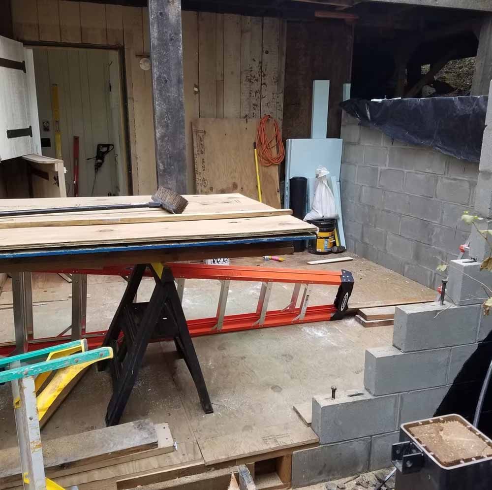 A cluttered workshop with a saw horse, stacked boards, cinder blocks, ladder, and wooden wall panels.