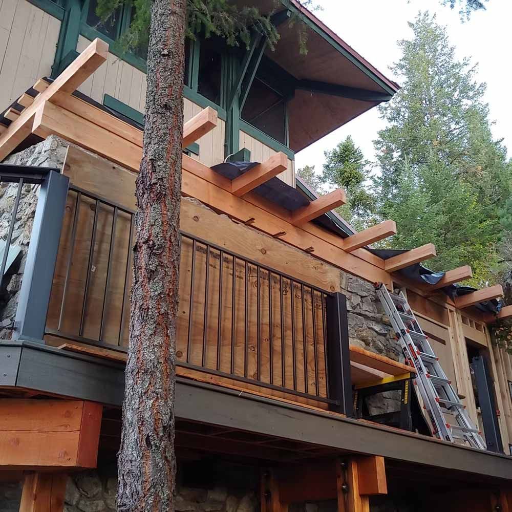 Wooden deck and balcony on a house under construction, with a ladder and surrounding trees.