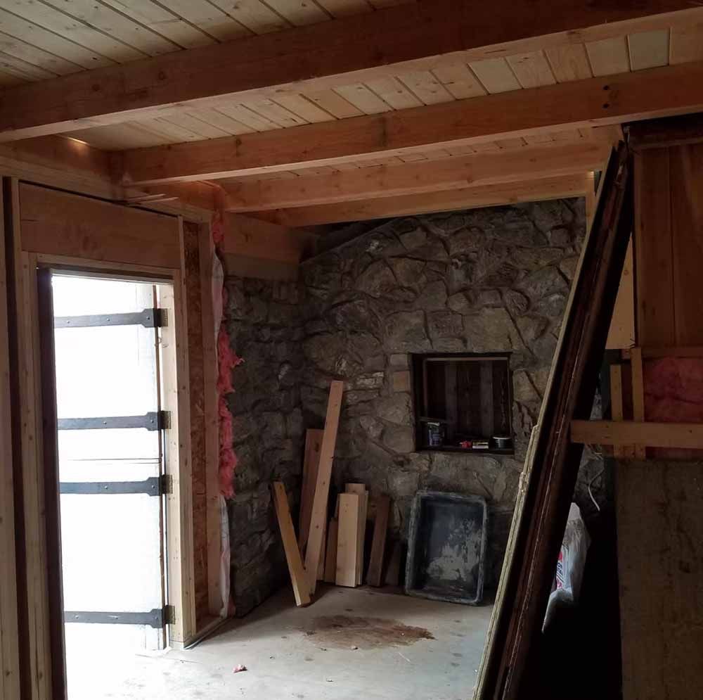 Unfinished room with stone fireplace, wooden beams, open door, and construction materials leaning against the wall.