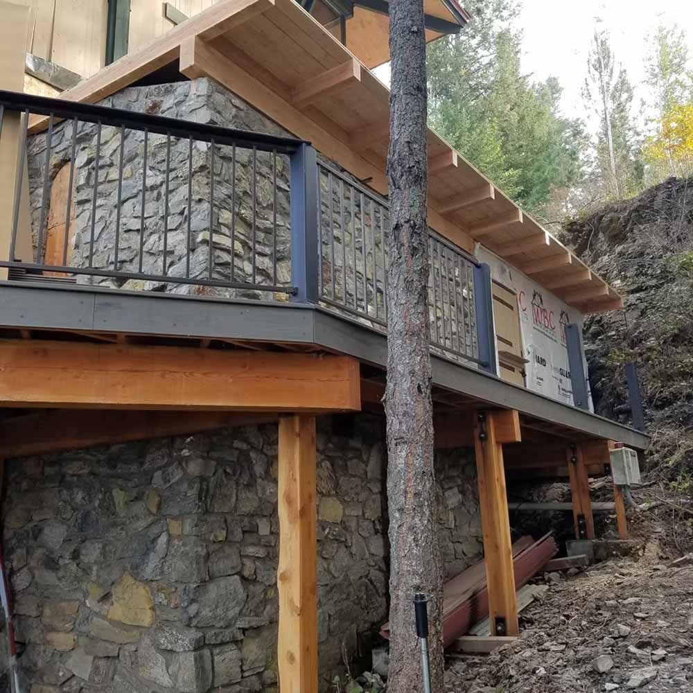Stone-and-wood house on a slope with a black metal balcony and exposed support posts.