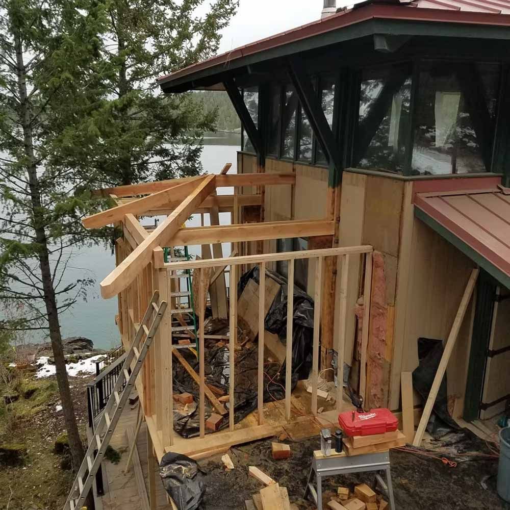 Wooden deck under construction beside a lakeside house, with exposed framing and tools.