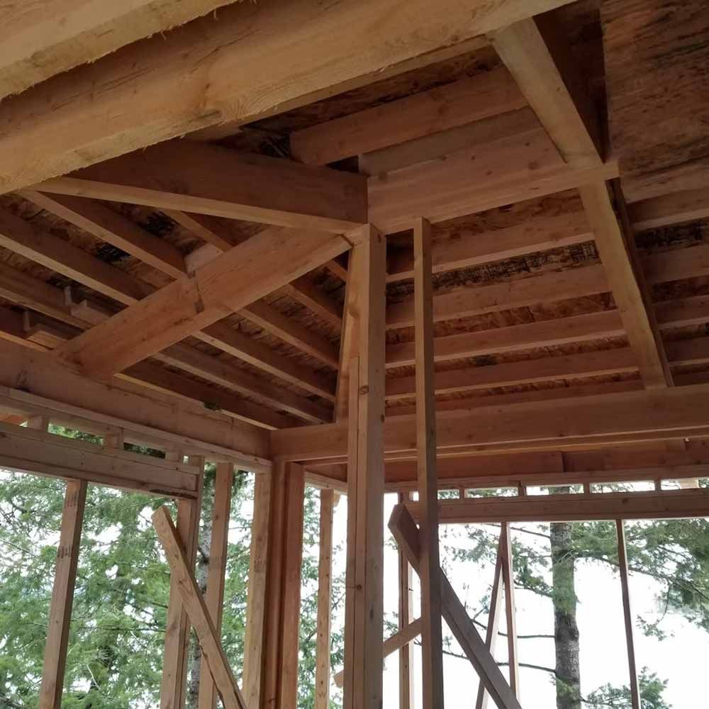 Wooden cabin roof framing under construction, viewed from inside with open wall framing and trees outside