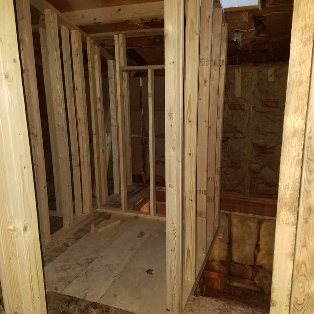 Unfinished wooden room framing with a narrow hallway and exposed studs on plywood floor.