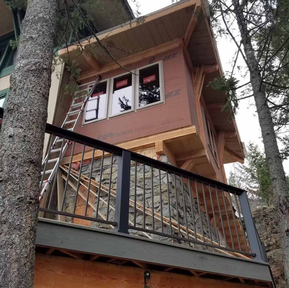 Treehouse-style wooden cabin with black railing, windows, and surrounding tall trees