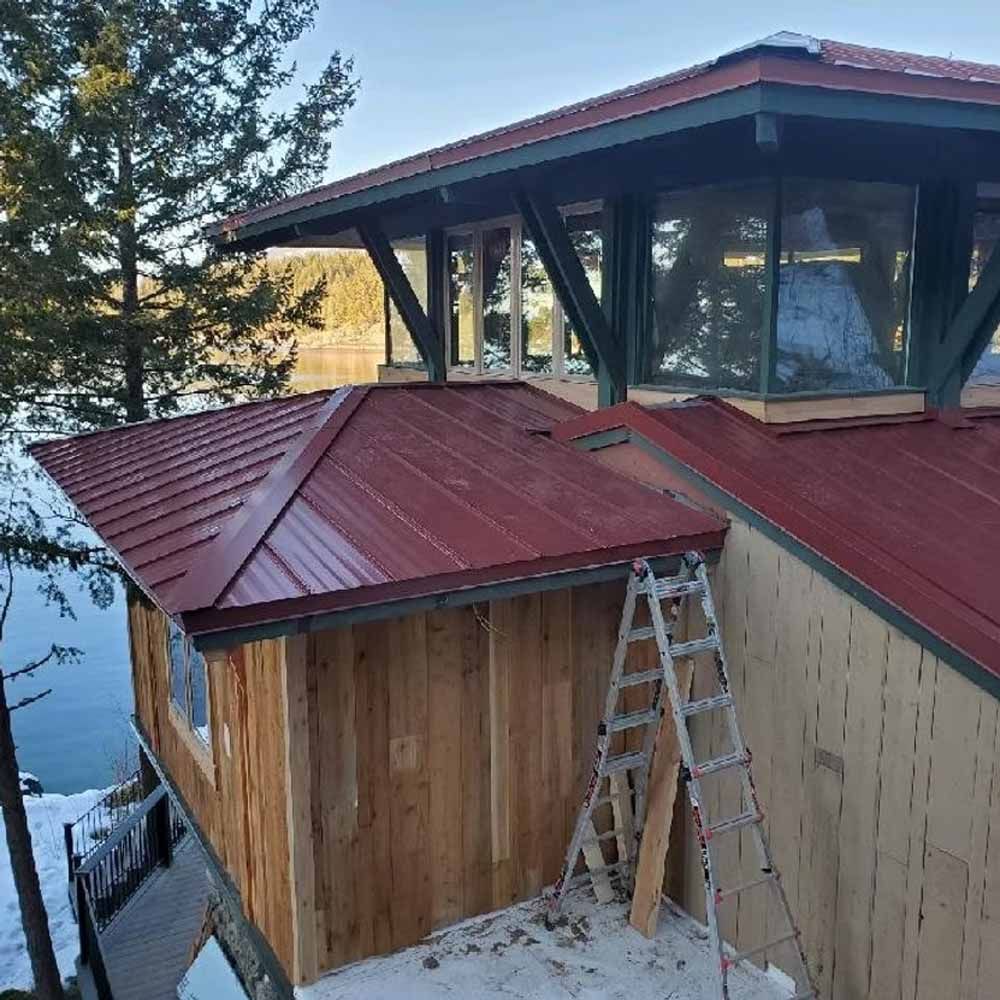 Alpine cabin with red metal roof and wooden siding beside a snowy lakeside, with an extension under construction