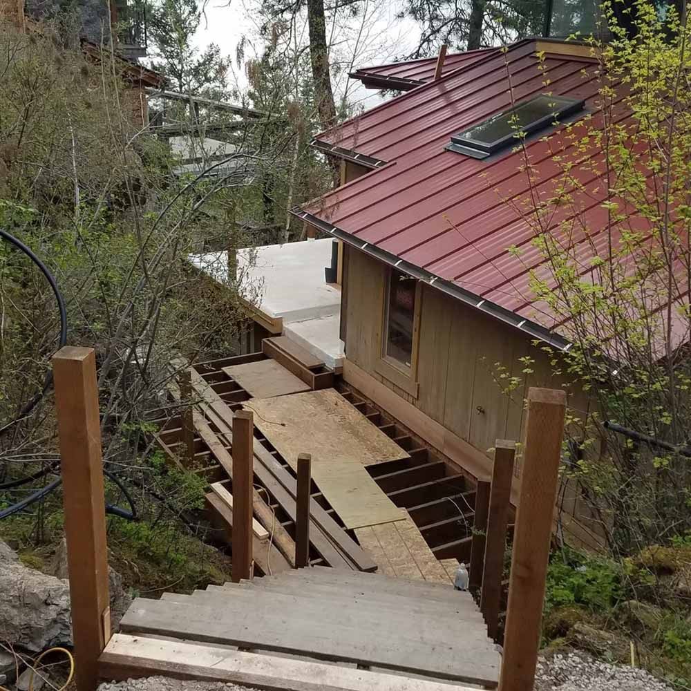 Wooden stairway leading to a beige house with a red metal roof on a wooded hillside.