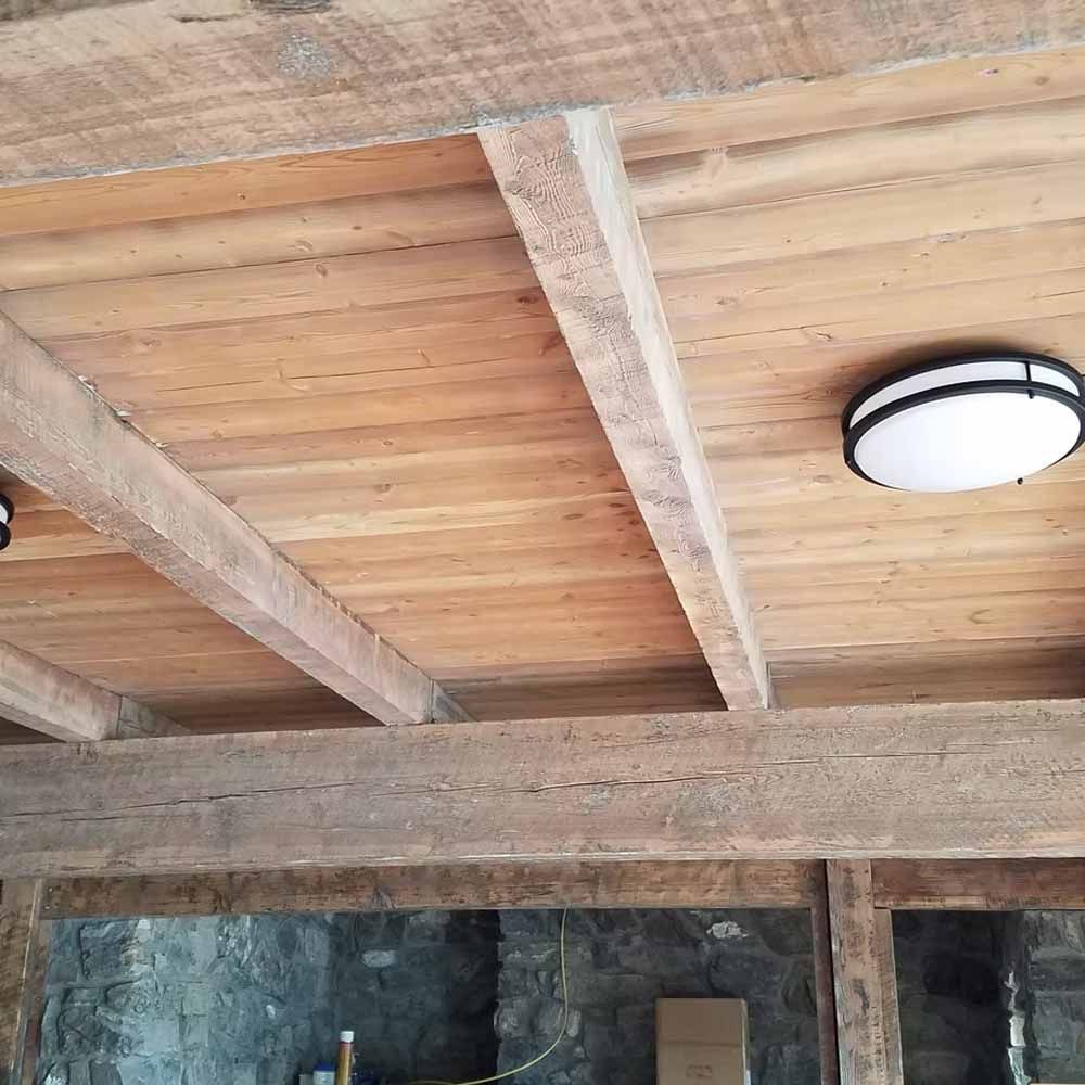 Wooden ceiling with exposed beams and a round flush-mount light above a stone wall.