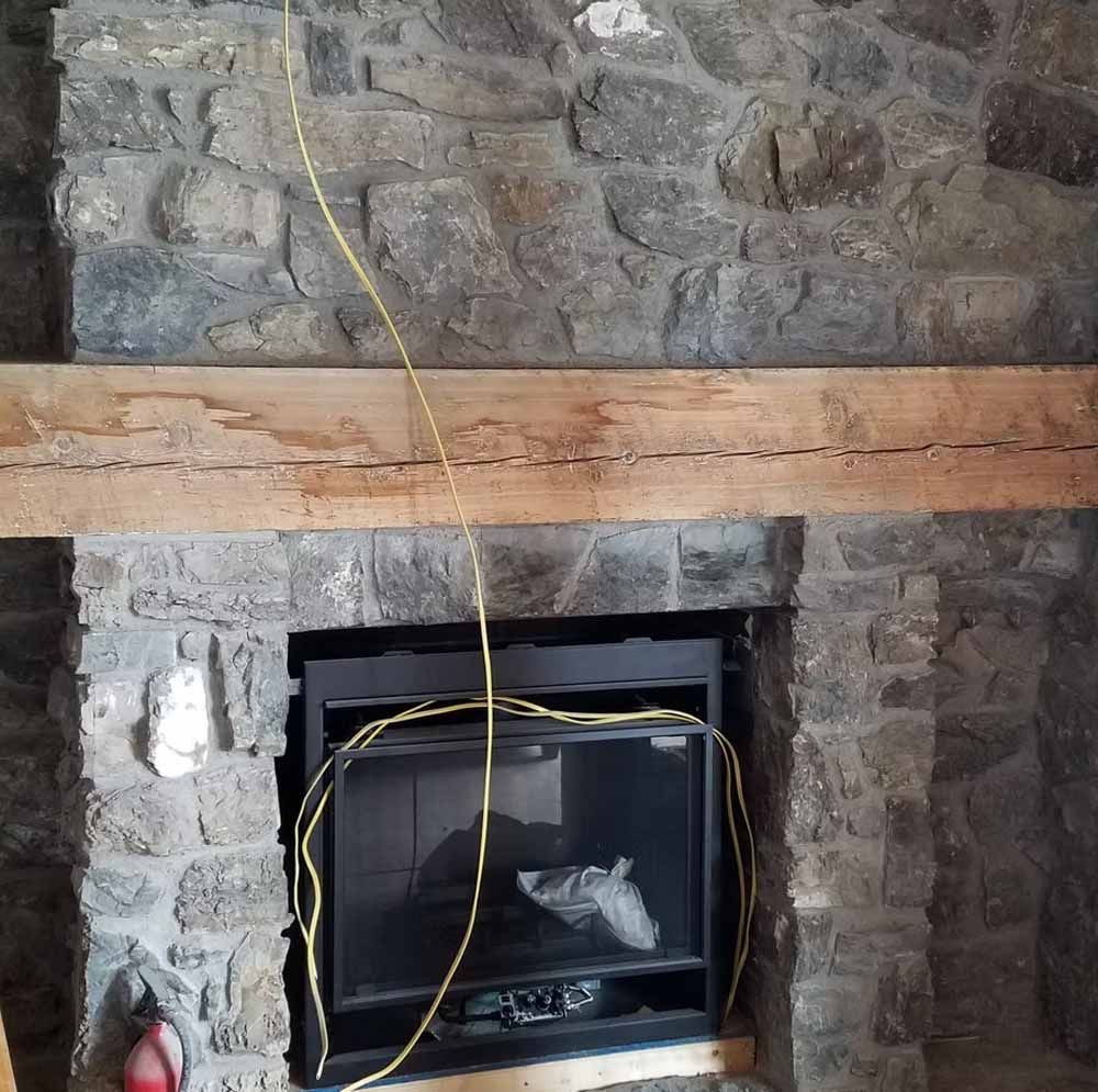 Stone fireplace with a wooden mantel and a black inset opening, with yellow cord hanging down.