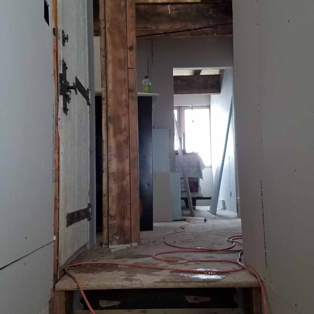 Narrow unfinished hallway with exposed wood framing, white walls, and construction debris on the floor