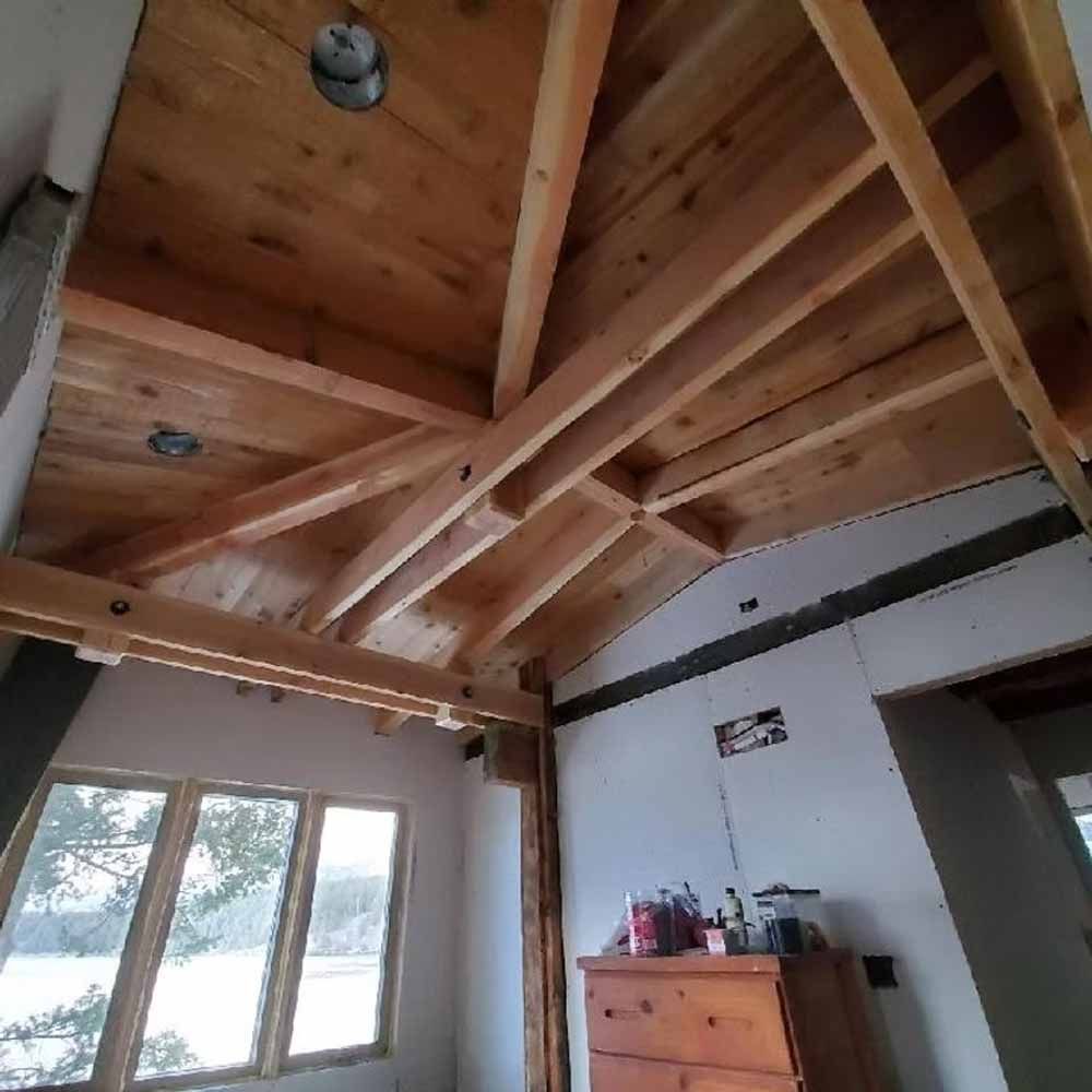 Wooden vaulted ceiling under construction above a bright room with windows and a small dresser.