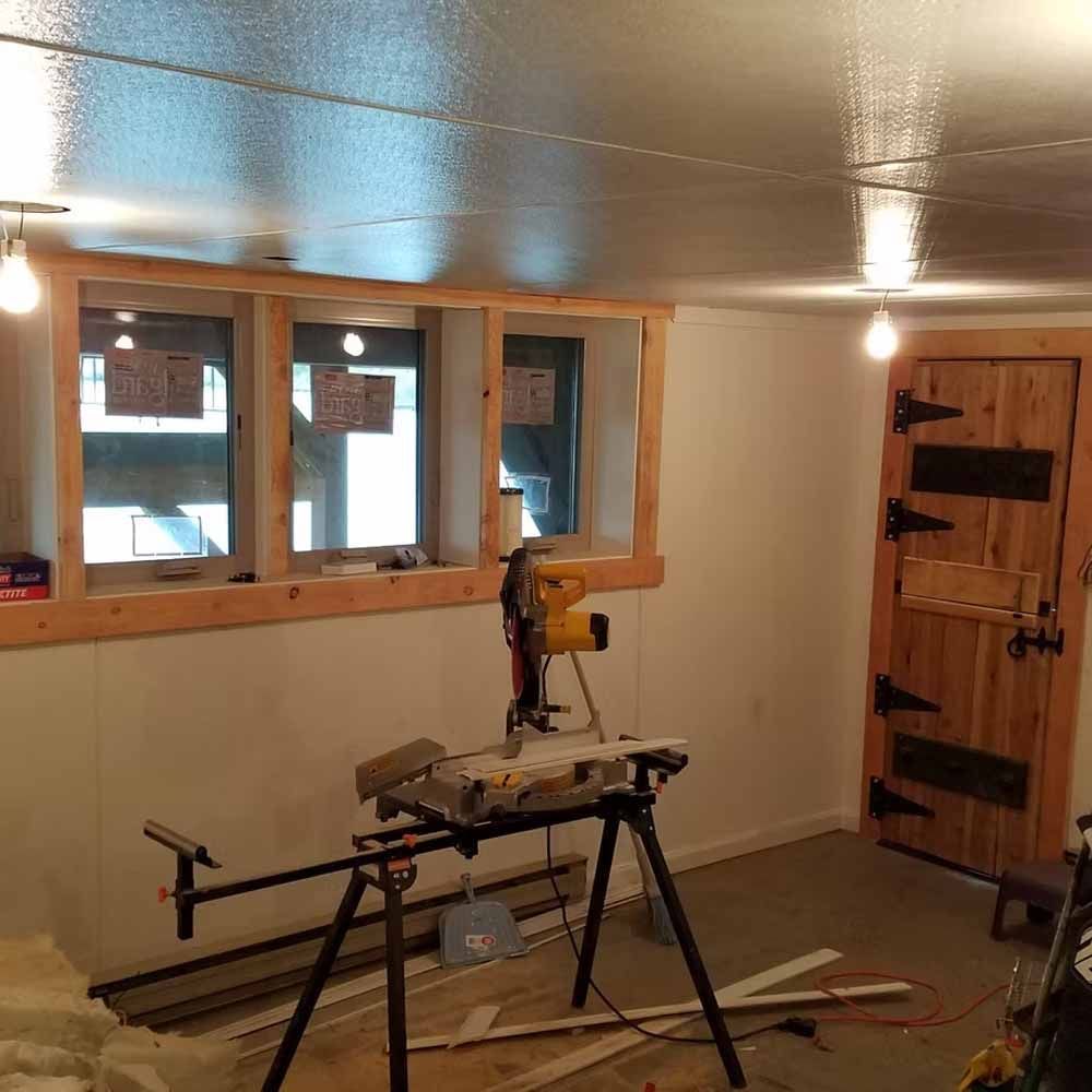 Small workshop with miter saw on stands, unfinished wood floor, windows, and a wooden door.