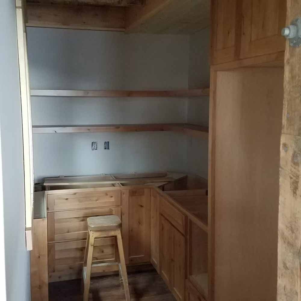 Empty unfinished kitchen nook with wooden cabinets, open shelves, and a stool in the corner.