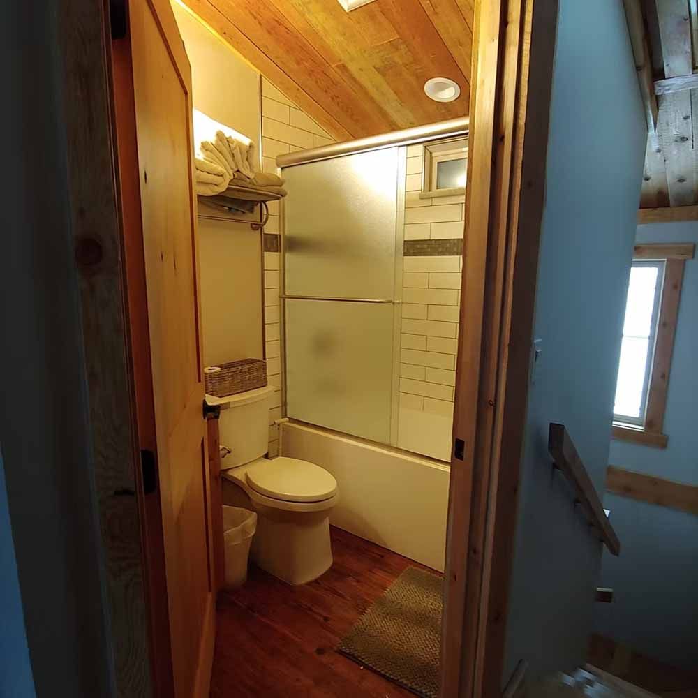 Small bathroom with toilet, shower, and wood floors, viewed through an open door