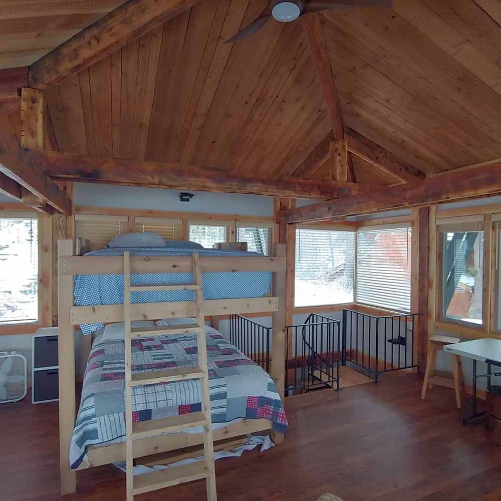 Loft bedroom with wooden A-frame ceiling, bunk bed, and large windows overlooking trees