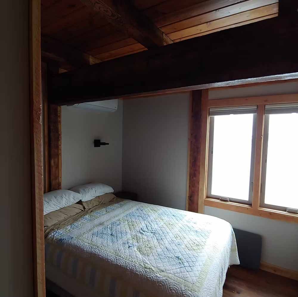 Small bedroom with a bed, wood ceiling beams, and two bright windows.